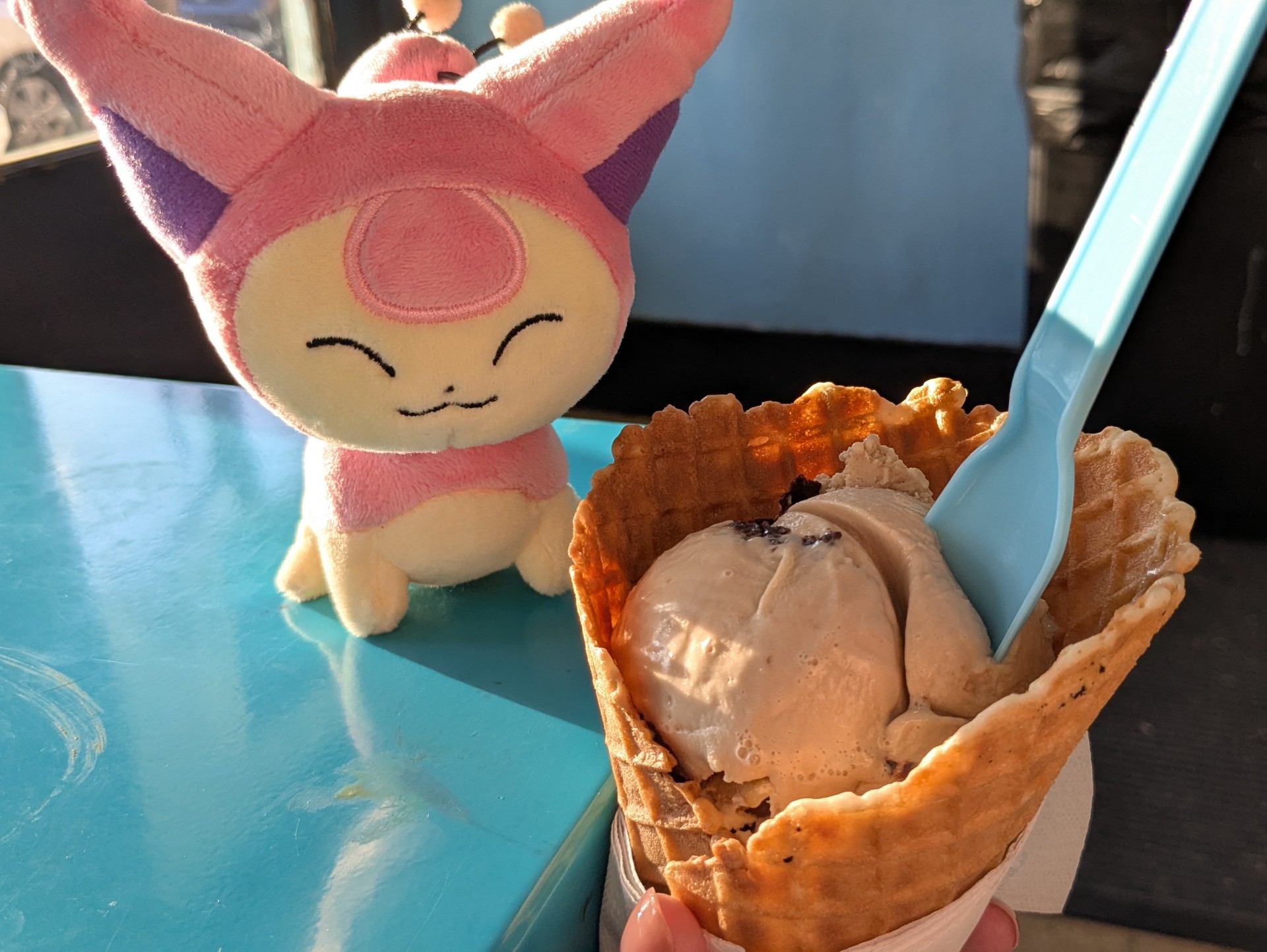 a scoop of ice cream in a waffle cone. a plush of a little pink cat Pokemon is sitting next to the ice cream.