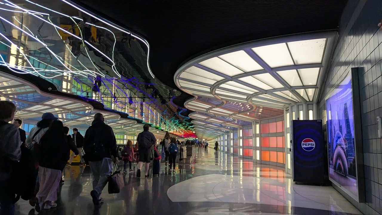 a hallway inside of an airport, with neon strands of light on the ceiling and glowing rainbow panels lining the walls